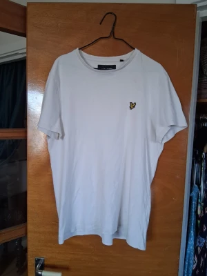 Vit t-shirt från Lyle & Scott - Vit t-shirt från Lyle & Scott med klassisk rund hals och korta ärmar. På bröstet finns den ikoniska gula fågel-loggan broderad. T-shirten är i mjuk bomull och har en clean och stilren look som passar till det mesta! 😃