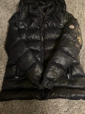 Svart dunjacka från Moncler med huva!  - Säl indicating en svart dunjacka från Moncler med huva och dragkedja framtill. Jackan har två fickor med dragkedja och Moncler-logga på ärmen. Puffig modell i glansigt material som håller dig varm under kalla dagar. Priset är diskuterbart 