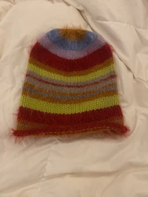 Randig fluffig mössa i mohairmix - Färgglad stickad mössa med breda ränder i rött, gult, lila, blått och orange. Mössan har en fluffig och mjuk känsla tack vare mohairmixen och har en avslappnad passform. Perfekt för dig som vill sticka ut med din stil.