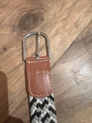 Randigt flätat bälte i svart och vitt - Snyggt flätat bälte med randigt mönster i svart och vitt. Spännet är i silverfärgad metall och ändarna är förstärkta med brunt skinn. Perfekt accessoar för att lyfta din outfit.