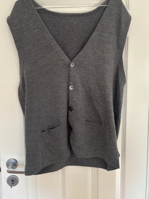 Grå stickad väst med knappar - Snygg grå stickad väst med djup V-ringning och fyra knappar framtill. Västen har två små fickor och är tillverkad i ett mjukt, tunt material. Perfekt att slänga över en skjorta eller t-shirt för en chill och stilren look.