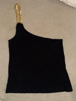Svart oneshoulder topp med guldkedja - Snygg svart oneshoulder topp med bred guldfärgad kedja som axelband. Toppen är ribbad och har en tight passform, perfekt för att sticka ut. Materialet känns mjukt och följsamt.
