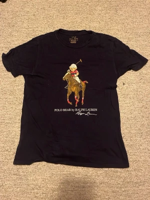 Mörkblå Polo Bear t-shirt från Ralph Lauren - Cool mörkblå t-shirt från Ralph Lauren med Polo Bear-tryck på bröstet där björnen rider häst och spelar polo. Klassisk rund hals och korta ärmar. Perfekt för dig som gillar streetstyle och ikoniska prints. Storlek 14-16 år men sitter som xs-S 