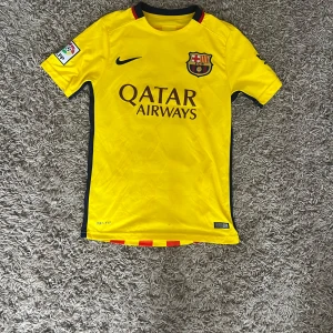 Barcelona Neymar JR 11 bortatröja Nike 2015 - Barcelona bortatröja med Neymar JR 11 på ryggen. Tröjan är gul med röda vertikala ränder på baksidan, svart Nike-logga och klubbmärke på bröstet. Qatar Airways-tryck på framsidan. Tillverkad i Dri-FIT-material för bästa komfort.