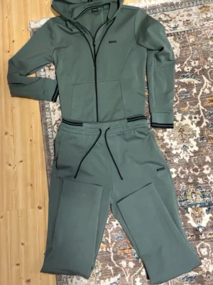 Grön loungewear-set från BOSS - Snyggt grönt loungewear-set från BOSS med matchande hoodie och byxor. Setet har svarta detaljer som dragkedja, snören och logga. Hoodien har fickor och byxorna har breda ben och resår i midjan. Perfekt för chill dagar hemma.