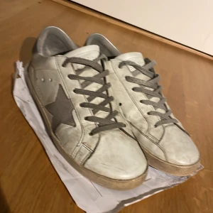 Golden Goose vita sneakers med stjärna - Golden Goose sneakers i vitt skinn med grå mockadetaljer och grå snörning. Sidan pryds av en ikonisk grå stjärna och baksidan har silvrig hälkappa med logga. Platt sula och rund tå ger en klassisk streetstil.