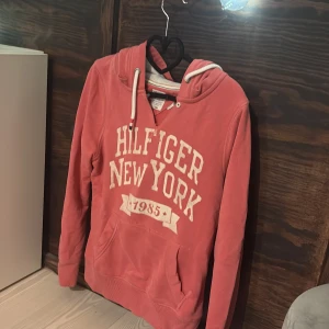 Coral färgad hoodie från Tommy Hilfiger  - Trendig snygg coral rosa hoodie från Tommy Hilfiger Denim med vit text 'Hilfiger New York 1985' på bröstet. Hoodien har huva med vita snören och känguruficka framtill. Perfekt för en avslappnad och trendig stil. Köpt begagnad men fortfarande väldig bra skick!