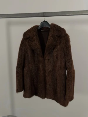 Brun äkta pälsjacka  - Vintage pälsjacka i äkta mink. Pälsen har en mörkbrun färg med tät, kort och mycket mjuk struktur samt naturlig glans därav troligen mink. Alternativt att det är bäver, vilket är en aning tåligare än mink.  Modellen är rak med klassisk krage och knäppning framtill med hyskor/hooks. Helfodrad insida i mörkbrunt tyg. Invändigt finns även en liten ficka med broderade initialer. Uppskattas passa XS–S beroende på önskad passform. Bra vintage-skick. Pälsen är mjuk och välbevarad och fodret är helt