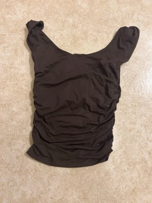 Brun ärmlös topp från Gina Tricot - Snygg brun ärmlös topp från Gina Tricot med rundad halsringning och rynkade sidor. Tillverkad i mjuk och stretchig polyesterblandning som sitter skönt mot kroppen. Perfekt att styla med jeans eller kjol för en trendig look. Helt ny inte använd 