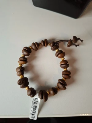 Armband med kaffebönor och pärlor - Unikt armband med bruna kaffebönor och små guldfärgade pärlor trädda på en brun tråd. Justerbar knytning för flexibel passform. Perfekt accessoar för dig som gillar naturliga material och en bohemisk stil.