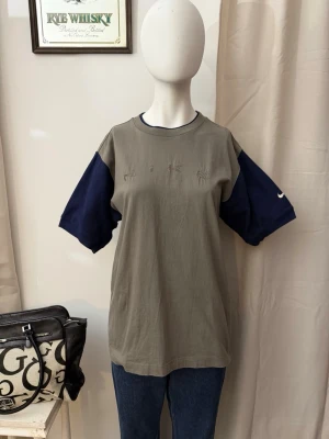 90’s Grå Nike t-shirt med blå ärmar - Cool t-shirt från Nike i grått med kontrasterande mörkblå korta ärmar. Framtill står NIKE broderat i samma färg som tröjan för en clean look. Perfekt till jeans eller shorts för en avslappnad stil.