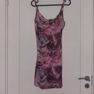 Mönstrad kort klänning med smala axelband - Säljer en kort klänning med smala axelband och ett unikt mönster i rosa, lila och svart. Klänningen har en figurnära passform och är perfekt för dig som gillar färg och att sticka ut. Materialet känns mjukt och stretchigt.