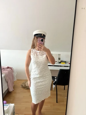 Studentklänning Vit spetsklänning  - Säljer en elegant vit klänning med spetsdetaljer och ärmlös form. Klänningen har en kroppsnära passning och en klassisk rund halsringning. Perfekt för student eller sommarens speciella tillfällen. Helt felfri!