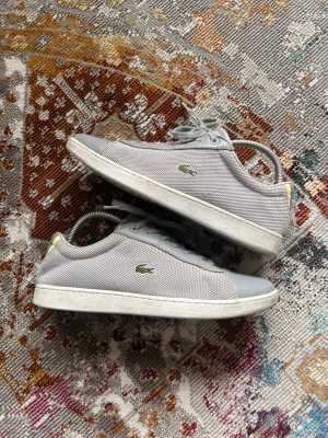 Gråa Lacoste sneakers i mesh och skinn - Snygga gråa sneakers från Lacoste med klassisk låg profil. Skorna har ovandel i mesh och detaljer i skinn, samt den ikoniska gröna krokodilloggan på sidan. Vita sulor och gråa skosnören ger en clean look. Perfekta för dig som gillar stilrena och sportiga skor.