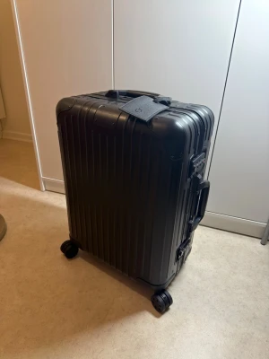 Svart kabinväska från Rimowa - Snygg svart resväska från Rimowa med hårt skal och fyra hjul för smidig rullning. Väskan har räfflad yta, integrerat TSA-lås och praktiska bärhandtag på både sidan och toppen. Perfekt för dig som vill resa med stil och funktion.