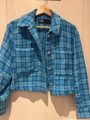 Blå kavaj från Zara - Snygg blå kavaj från Zara i tweedmaterial med rutigt mönster och silvriga knappar. Kavajen har krage, två bröstfickor och en croppad passform. Perfekt för att lyfta din outfit med en trendig touch.