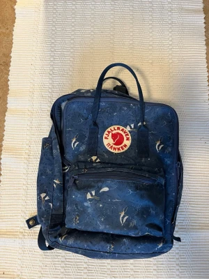 Blå Fjällräven Kånken ryggsäck mönstrad - Säljer en blå Fjällräven Kånken ryggsäck med unikt mönster i vitt och beige. Klassisk fyrkantig form, stort huvudfack, framficka med dragkedja och två sidofickor. Ikonisk röd logotyp framtill. Tillverkad i slitstarkt syntetmaterial.