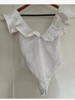 Vit volang body - Säljer en vit body med djup v-ringning och stora volanger vid axlarna. Helt oanvänd!!