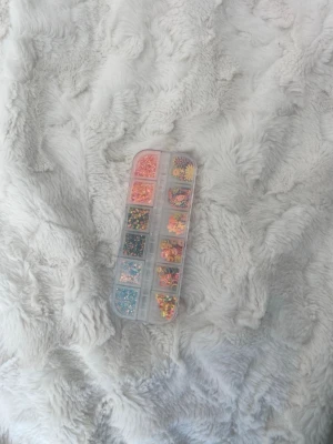 Nageldekorationer i mixade färger - Säljer ett set med nageldekorationer i olika former och färger, perfekt för att skapa kreativa och unika nageldesigner. Dekorationerna kommer i en praktisk plastlåda med flera fack och innehåller bland annat stjärnor, blommor och glitter i pastell och metallic.