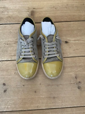 Gråa  Lanvins sneakers med gula detaljer - Säljer ett par gråa Lanvins sneakers med gula detaljer runt tå och kant. Skorna har klassisk låg profil, snörning och vit sula. Materialet är mocka och de har en stilren design som passar till jeans och t-shirt.