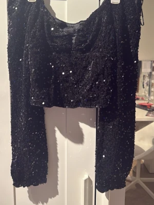 Svart glittrig croppad topp - Säljer en svart croppad topp med långa ärmar och massor av glittrande paljetter. Toppen har en snygg, åtsittande passform och är perfekt för dig som vill sticka ut. Materialet känns mjukt och stretchigt, och toppen har en trendig look med sin glittriga yta.