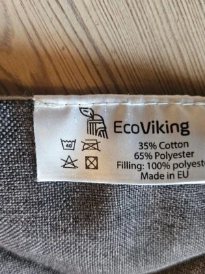 EcoViking grå vadderad kudde - Grå vadderad kudde från EcoViking med yttertyg i bomull och polyester samt fyllning av polyester. Kudden har en snygg brun detalj i konstläder med präglad logga. Passar perfekt som sitt- eller ryggstöd.