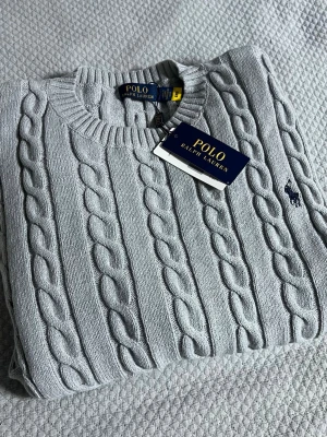 Ljusgrå kabelstickad tröja Polo Ralph Lauren - Snygg ljusgrå kabelstickad tröja från Polo Ralph Lauren med klassisk rund halsringning och mörkblå broderad logga på bröstet. Tröjan har en tidlös design och är tillverkad i mjukt material som passar perfekt till alla tillfällen.