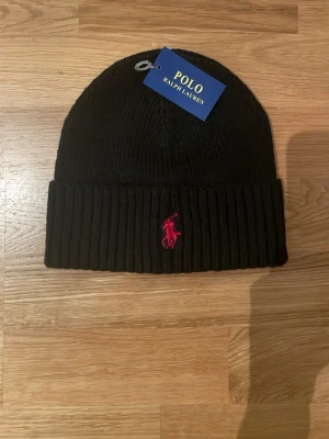 Svart ribbad mössa med Polo Ralph Lauren logga  - Snygg svart ribbstickad mössa med Polo Ralph Lauren logga broderad framtill. Perfekt accessoar för kalla dagar och enkel att matcha med olika outfits.
