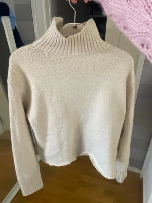 Beige stickad polotröja - Mysig beige stickad polotröja med hög krage och långa ärmar. Tröjan har en rak passform och breda ribbstickade muddar vid ärmslut och hals. Perfekt för kalla dagar och enkel att matcha med jeans eller kjol.