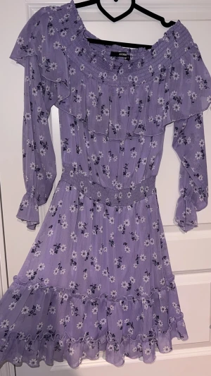 Lila off shoulder klänning  - En långärmad lila off shoulder klänning med blommor och volanger. Den har resår i midjan och vid handleden. Även en underklänning så den är inte genomskinlig. 