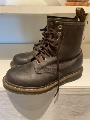 Bruna Dr. Martens boots i läder - Klassiska bruna Dr. Martens boots i äkta läder med snörning och gul söm runt sulan. Högt skaft, robust design och dragflik baktill med logga. Perfekta för dig som gillar en tidlös och edgy stil.