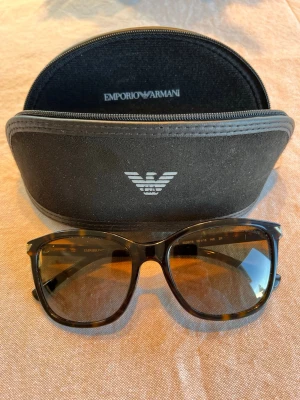 Bruna solglasögon från Emporio Armani - Snygga solglasögon från Emporio Armani med klassisk brunmelerad båge och bruna glas. Bågen har en fyrkantig form och logotypen syns på sidan. Medföljer originalfodral i svart. Inga repor. 
