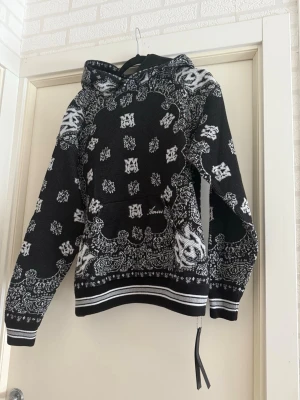 Svart och vit hoodie från Amiri (bandana) - Snygg svart hoodie från Amiri med vit paisley- och logomönster över hela tröjan. Stickad känsla, ribbade muddar och nederkant med vita ränder. Stor huva och cool streetwear-vibe. Perfekt för dig som gillar unika detaljer och trendig design.
