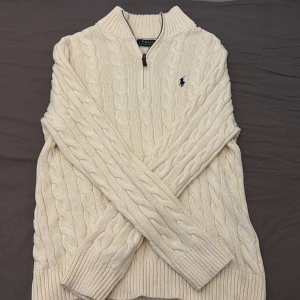 Beige stickad tröja Polo Ralph Lauren - Kabelstickad vit tröja från Polo Ralph Lauren med halv dragkedja och broderad marinblå logga på bröstet. Tröjan har justerbar krage och långa ärmar, perfekt för lager-på-lager under kyliga dagar. Klassisk och tidlös design i mjuk bomull. Nypris: 2900