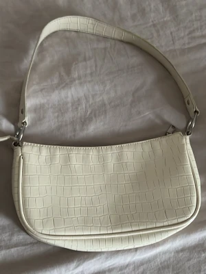 Vit axelväska med krokopräglat mönster - Stilren axelväska i vitt konstläder med krokopräglat mönster. Väskan har en kort axelrem och silverfärgade metalldetaljer. Perfekt för dig som gillar minimalistisk och trendig design.