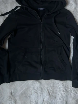 Svart zip hoodie med huva - Klassisk svart hoodie med dragkedja från Fruit of the Loom. Hoodien har justerbar huva med snören, två fickor framtill och ribbade muddar vid ärmar och nederkant. Perfekt för en avslappnad och stilren look.