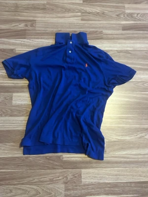 Blå pikétröja från Polo Ralph Lauren - Klassisk blå pikétröja från Polo Ralph Lauren med korta ärmar och krage. Tröjan har knappar framtill och den ikoniska lilla röda loggan broderad på bröstet. Perfekt för en stilren och avslappnad look.