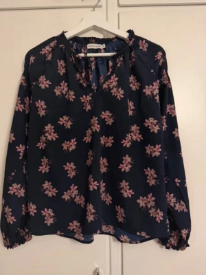 Blommig blus  - Mörkblå blus från Angelology, lager157 med rosa blommönster och v-ringning med knytband. Långärmad modell med resår vid ärmslut. Tunn och luftig kvalitet, perfekt för en trendig och avslappnad look.