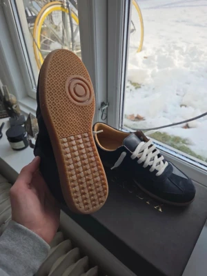 Masion Margiela - Stilrena svarta sneakers med vit detalj och vita snören. Skorna har en klassisk låg siluett och en brun gummisula med rutmönster undertill. Ovandelen verkar vara i mocka eller nubuck och insidan är ljusbrun. Perfekta för en clean och avslappnad look. Skorna e från Skorlyx insperirad av masion margiela nu prisa 1600 