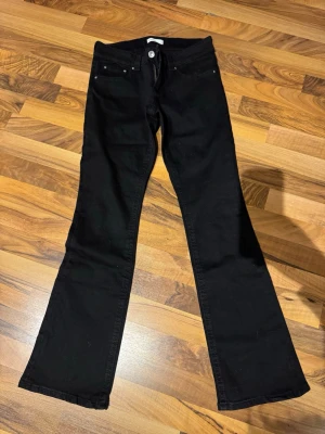 Svarta bootcut jeans - Svarta jeans med bootcut passform och klassisk femficksdesign. Jeansen har vida ben nertill och silverfärgade knappar och nitar. Perfekta för dig som gillar en retrostil med lite extra attityd.