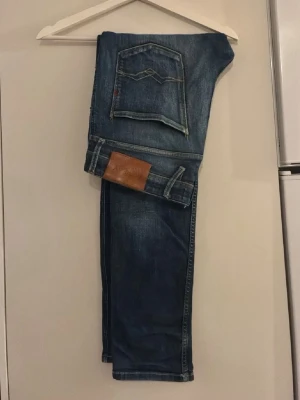 Blå jeans från replay Jeans - Säljer ett par klassiska blå jeans från Nudie Jeans med snygga slitningar och kontrastsömmar. Jeansen har fem fickor, rak passform och en brun läderpatch med logga bak i midjan. Perfekta för en avslappnad stil.
