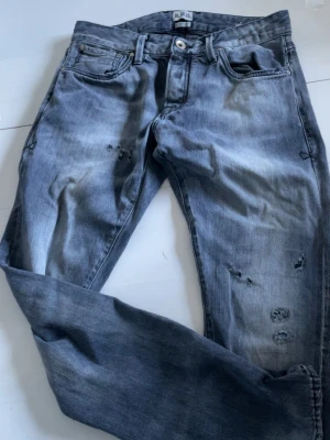 Grå slitna jeans från R.D.D. - Snygga grå jeans från R.D.D. med slitningar och distressed detaljer på både fram- och baksida. Klassisk femficksmodell med raka ben och knappgylf. Perfekta för en avslappnad och trendig look.