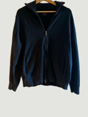 Marinblå ziptröja från Massimo Dutti - Stilren marinblå ziptröja från Massimo Dutti med hög krage och tvåvägsdragkedja. Tröjan har långa ärmar, ribbade muddar och två fickor framtill. Perfekt för lager-på-lager och enkel att matcha med olika outfits.