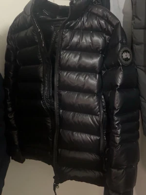 Svart dunjacka från Canada Goose - Snygg svart dunjacka från Canada Goose med glansig finish och klassisk patch på ärmen. Jackan har hög krage, dragkedja framtill och breda, horisontella kanaler fyllda med dun. Perfekt för kalla vinterdagar och riktigt varm.