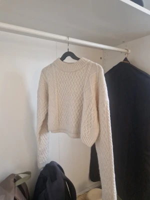 Beige croppad stickad tröja - Säljer en beige croppad stickad tröja med grovt kabelstickat mönster framtill och på ärmarna. Tröjan har rund halsringning och ribbade muddar. Perfekt för lager på lager och en trendig look.