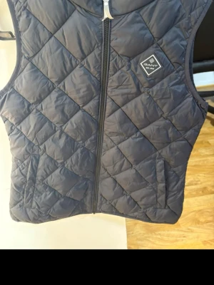 Mörkblå dunväst från GANT - Snygg mörkblå dunväst från GANT med quiltat mönster och dragkedja framtill. Västen har två fickor och en diskret logotyp på bröstet. Perfekt för lager-på-lager och ger en clean look till din outfit.