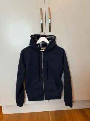 Burberry Hoodie Strl S/XS - En Marinblå Hoodie i storlek S men den passar bättre i storlek XS. Den är inte äkta därav priset men den är i bra skick. Det klassiska Burberry mönstret på insidan av hoodien gör den till en unik design. Säljer den eftersom den är för liten.