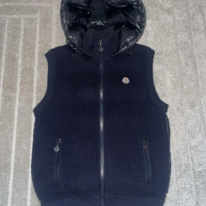 Mörkblå stickad väst med dunhuva - Snygg mörkblå stickad väst från Moncler med en quiltad dunhuva. Västen har dragkedja framtill och två praktiska fickor med dragkedjor. Perfekt för kyligare dagar.