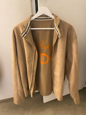 Beige jacka från Reporter RDG Milano - Snygg beige jacka från Reporter RDG Milano med klassisk krage och diskreta ränder på insidan av kragen. Jackan har dragkedja framtill och två fickor. Materialet känns mjukt och har en mockaliknande yta, perfekt för dig som gillar stilrena och tidlösa plagg.