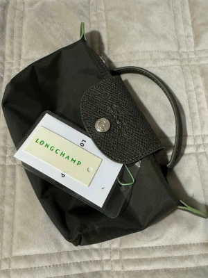 Mörkgrön handväska från Longchamp - Snygg mörkgrön handväska från Longchamp med klassisk vikbar design. Väskan har ett lock i präglat läder med silverfärgad knapp och handtag i matchande läder. Tillverkad i slitstark nylon med gröna detaljer på sidorna. Perfekt för dig som gillar stilrena accessoarer.